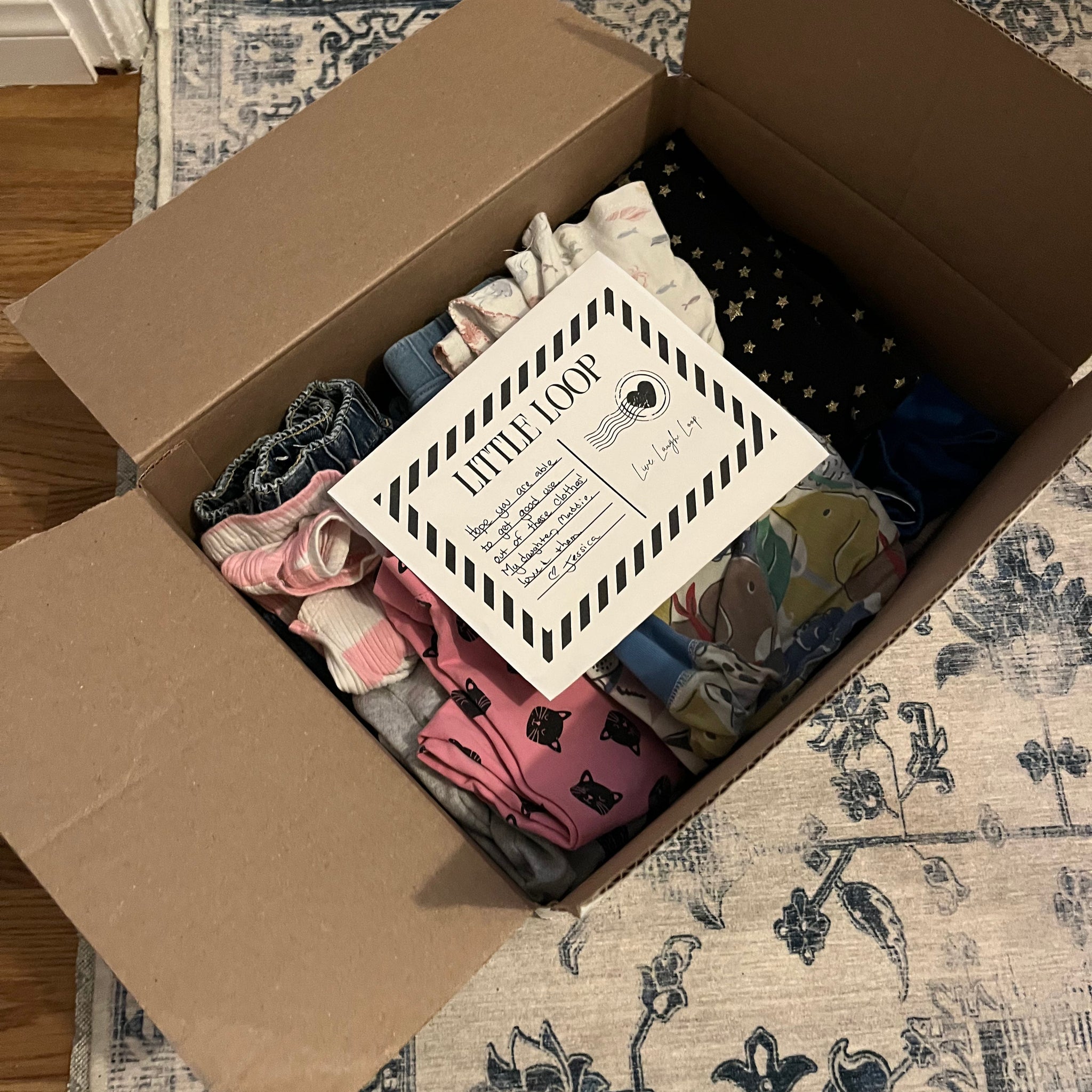 A cardboard box filled with folded baby clothes, a handwritten note tucked inside, and a smiling parent ready to tape it shut—part of a clothing swap system for parents.