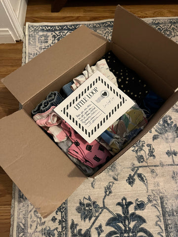 A cardboard box filled with folded baby clothes, a handwritten note tucked inside, and a smiling parent ready to tape it shut—part of a clothing swap system for parents.