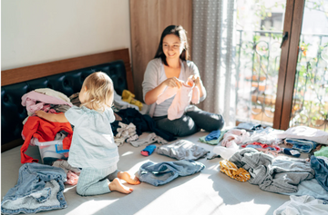A parent kneels on the floor sorting toddler clothes into labeled donation, storage, and keep bins in a bright, organized room.