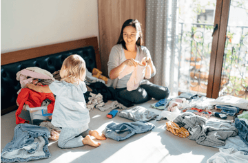 A parent kneels on the floor sorting toddler clothes into labeled donation, storage, and keep bins in a bright, organized room.
